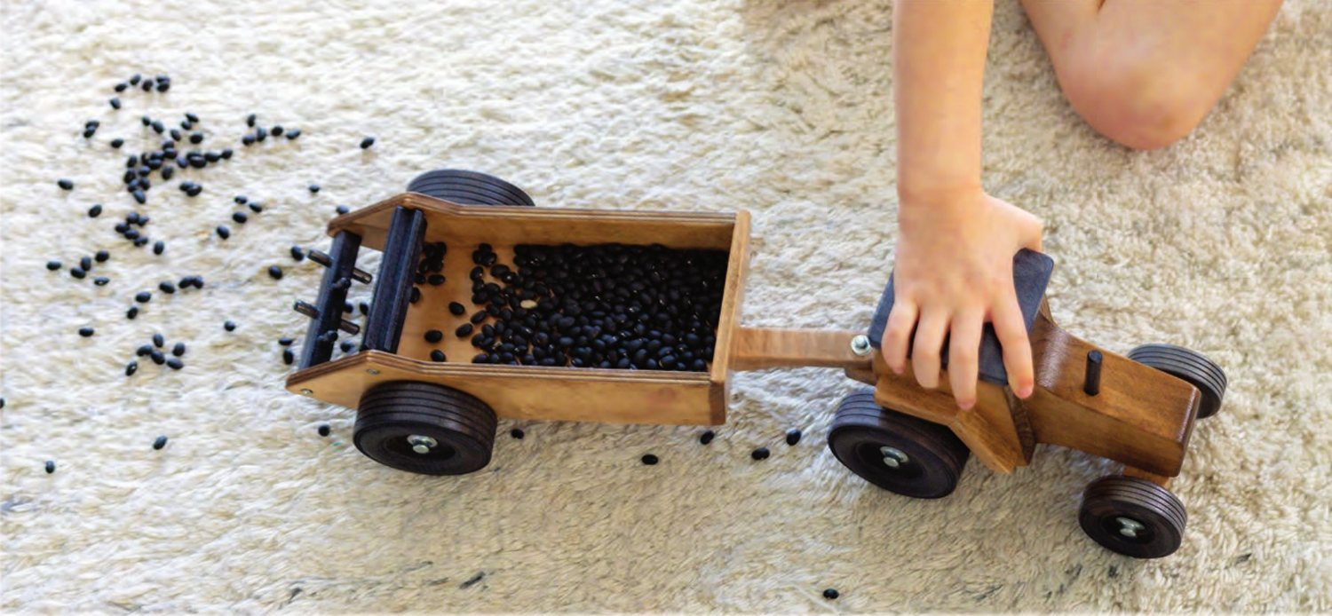 Child's hand pulling a tractor and manure spreader loaded with black beans across a rug