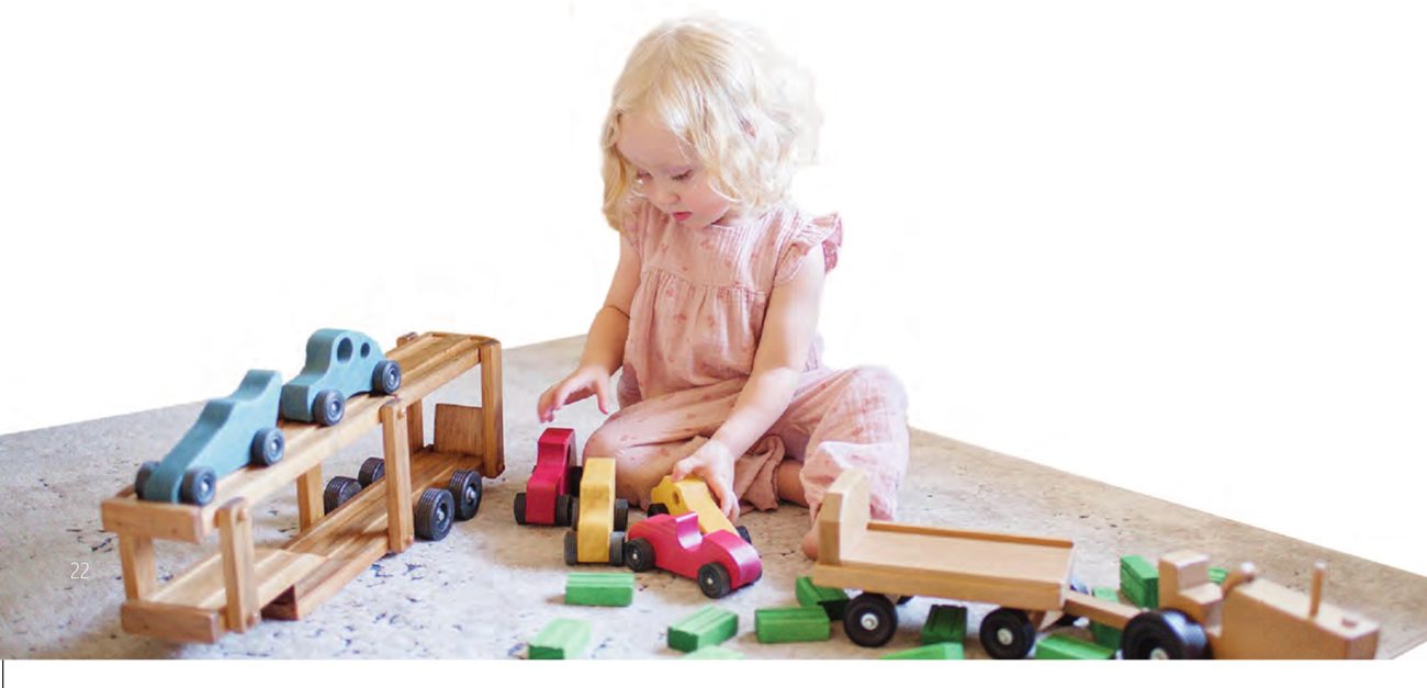 Little girl playing with a wooden car carrier and colorful wooden cars on a rug